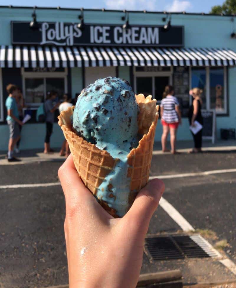 Hand holding a waffle cone with blue ice cream and chocolate chips in front of an ice cream shop with a striped awning. People are lined up outside.