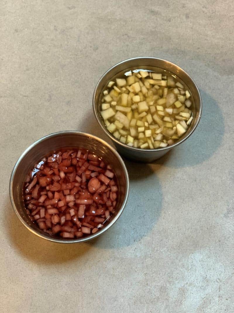 Two metal bowls contain finely chopped apples soaking in liquid; the left liquid is reddish, and the right is yellowish.