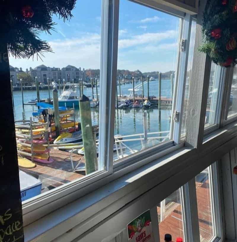 View through an open window showing a marina with boats docked at wooden piers, colorful paddleboards stacked nearby, and buildings across the water under a clear blue sky.