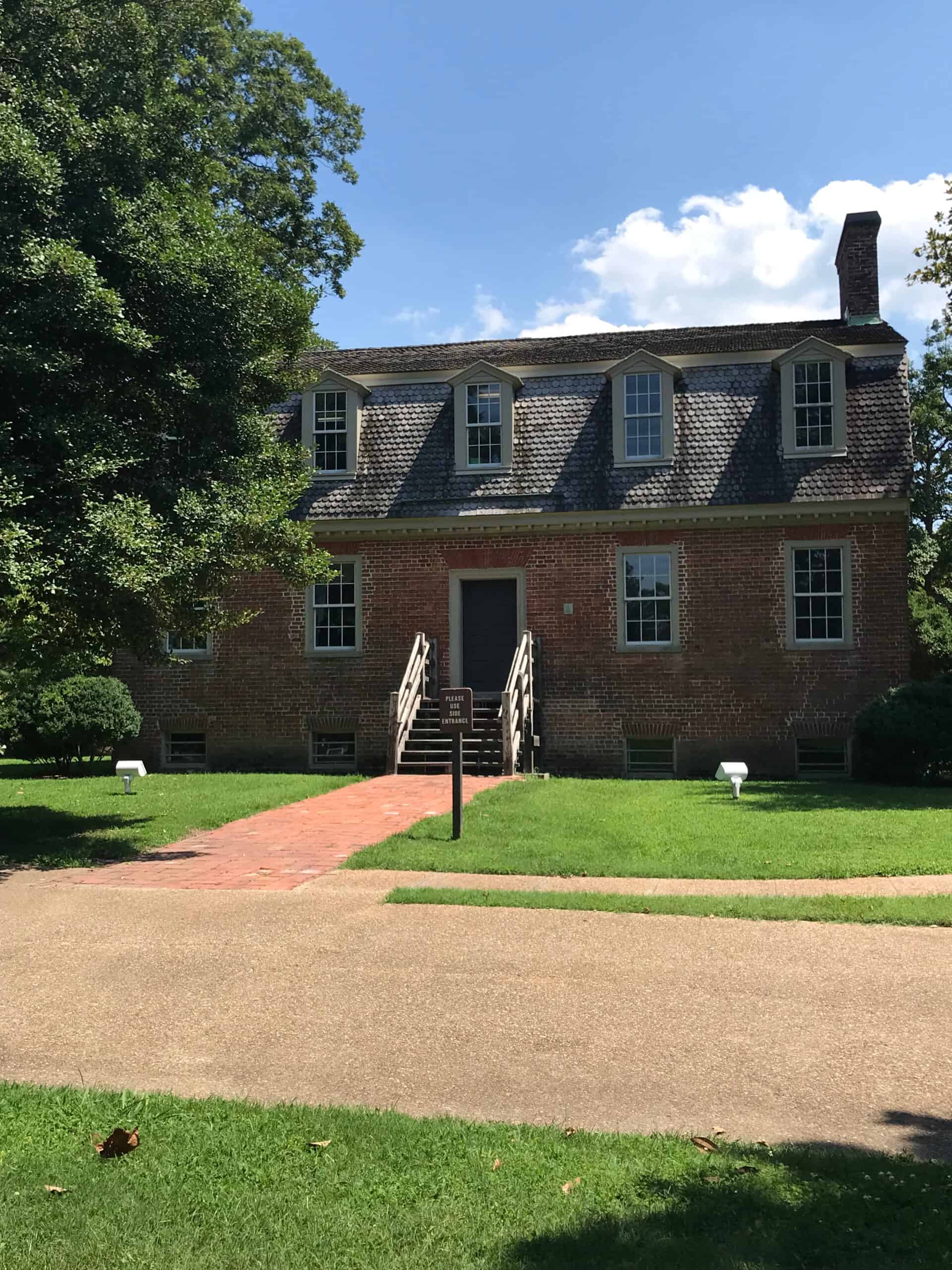 A brick colonial-style house with a gambrel roof, central entrance with stairs, and manicured lawn, under a partly cloudy sky.