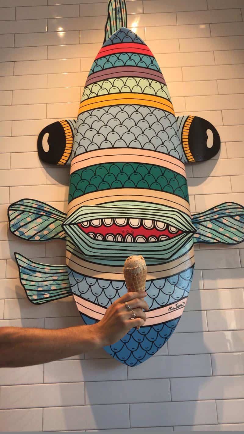A hand holds an ice cream cone in front of a colorful, striped fish art piece mounted on a tiled wall.