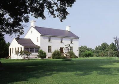A white, two-story house with a dark roof sits on a grassy lawn surrounded by trees under a clear sky.