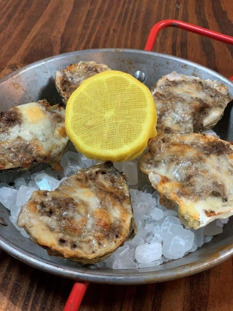 Five baked oysters with a yellow lemon slice on top, served on a bed of ice in a metal dish with a red handle.