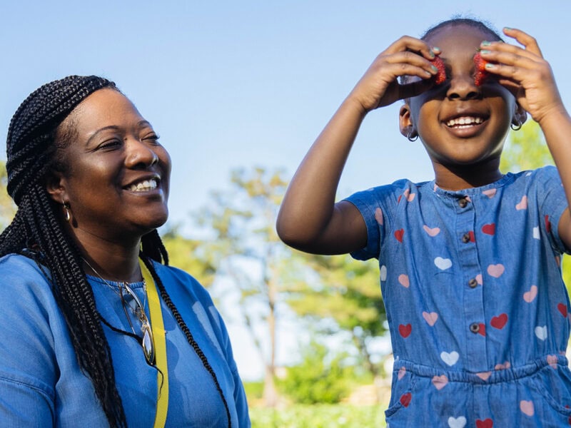 A smiling woman watches as a girl playfully holds strawberries to her eyes in a sunny outdoor farm area.