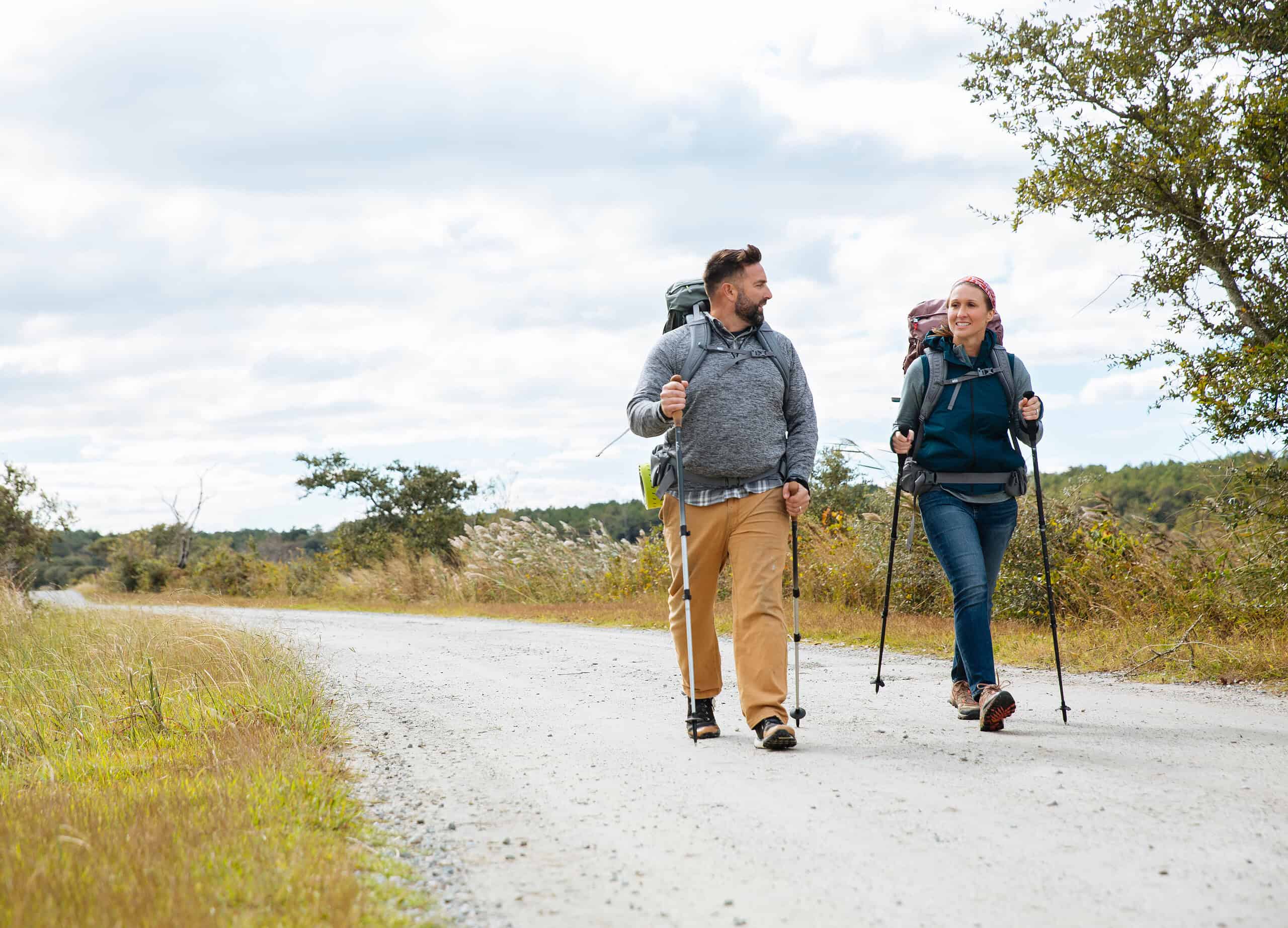 Two people hiking with backpacks and walking poles on a gravel path surrounded by fields and shrubs under a cloudy sky.