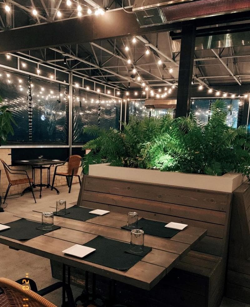 Outdoor restaurant seating area with wooden tables, chairs, string lights, and a planter with ferns, under a covered patio.
