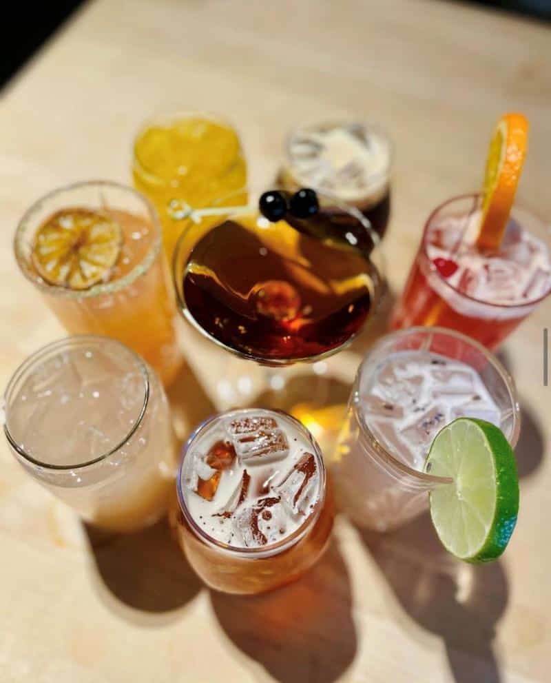 A top view of various cocktails arranged on a table. Drinks are garnished with lime, orange slices, cherries, and herbs. Glasses contain ice and colorful liquids.