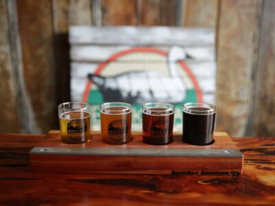 A wooden flight board holds four glasses of beer, varying in color from light to dark. A logo is visible on each glass, and a blurred background features a rustic sign with a goose illustration.