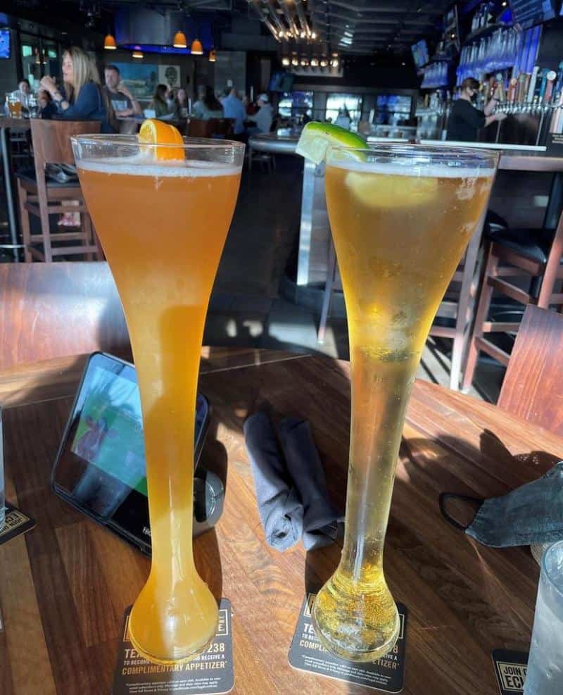 Two tall, curvy beer glasses on a wooden table in a bar. One has an orange wedge; the other has a lime. People sit at tables in the background.