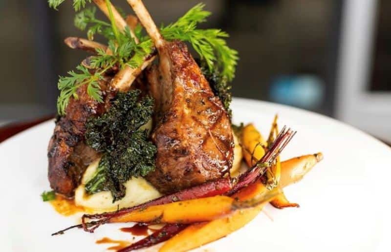 Grilled lamb chops on mashed potatoes with roasted carrots and greens, garnished with fresh herbs on a white plate.