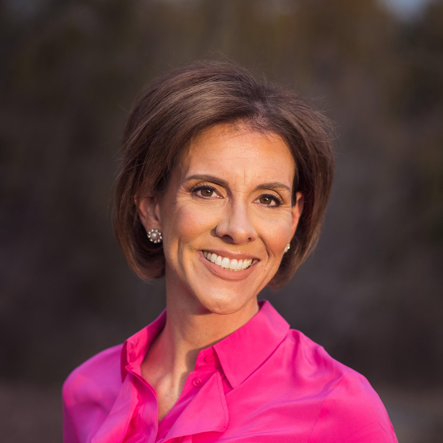 Author A woman with short brown hair, wearing a bright pink button-up shirt and pearl earrings, smiles outdoors with a blurred background.