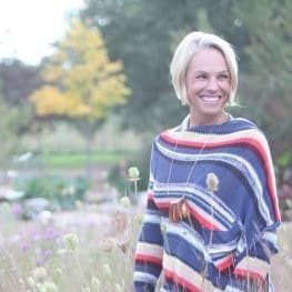 Author Staci Amend is a Denver based creative writer, smiling wearing a colorful striped poncho stands in a field of wildflowers, surrounded by greenery and autumn foliage.