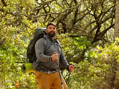 Man hiking on a forest trail with a backpack and trekking poles, surrounded by lush green trees and vegetation.