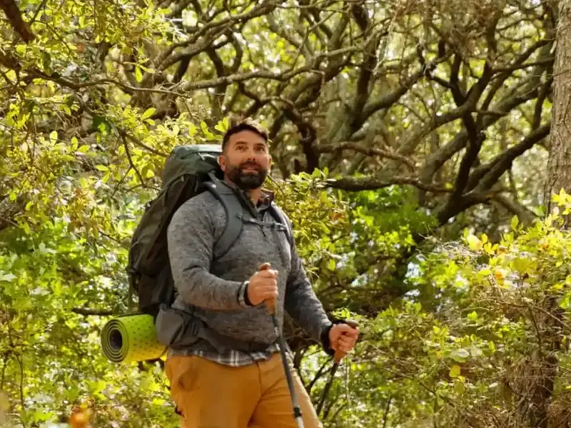 Man hiking on a forest trail with a backpack and trekking poles, surrounded by lush green trees and vegetation.