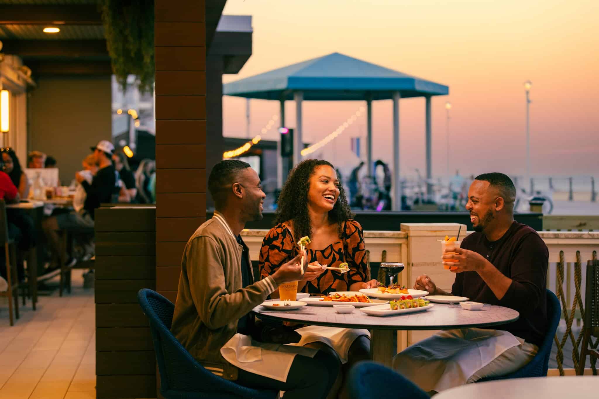 Three people sit at an outdoor restaurant table, smiling and talking while enjoying food and drinks at sunset.