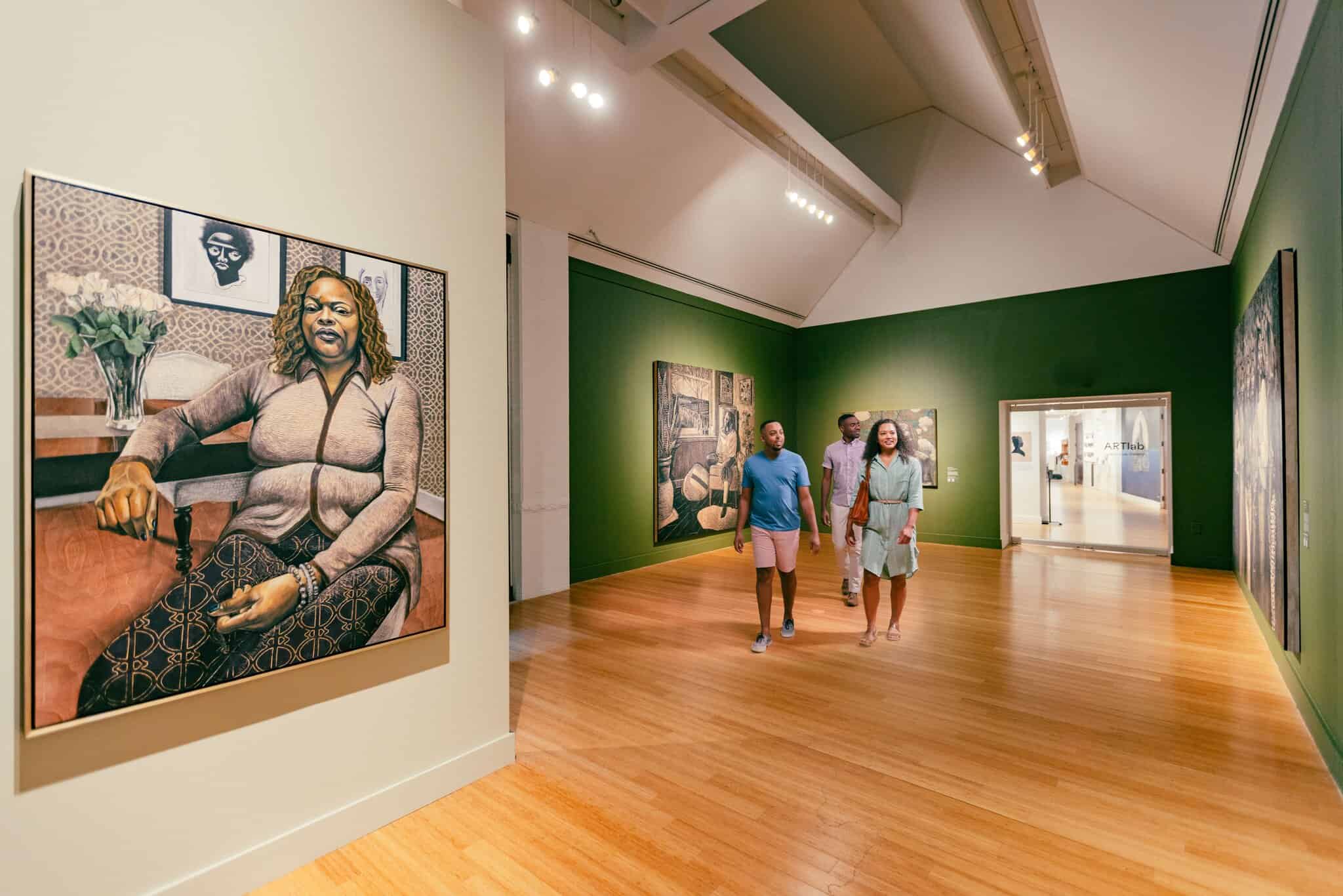 Three people walk through the Virginia Museum of Contemporary Art, with green walls, observing large portrait paintings displayed around the room.