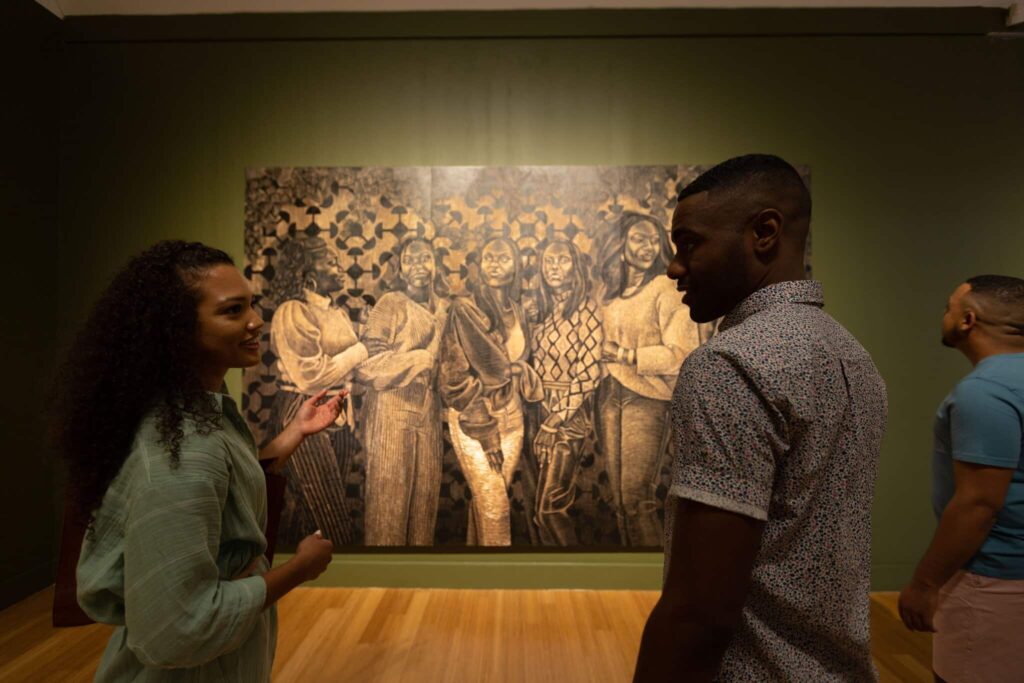 Three people view a large, detailed painting of a group of figures in the Virginia Museum of Contemporary Art with green walls and wooden floors.