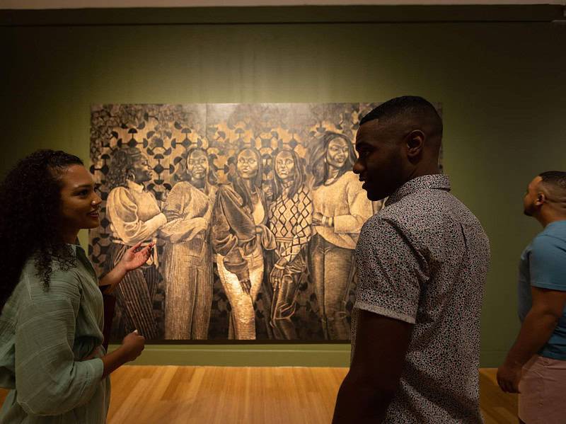 Three people view a large, detailed painting of a group of figures in the Virginia Museum of Contemporary Art with green walls and wooden floors.