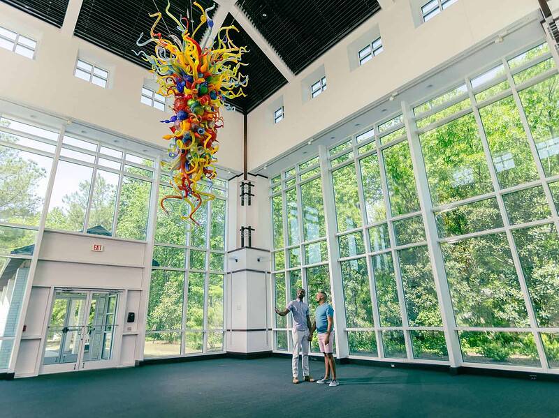 Two people stand in a spacious, sunlit room with large windows, looking up at a colorful glass sculpture by Dale Chihuly hanging from the ceiling at the Virginia Museum of Contemporary Art.