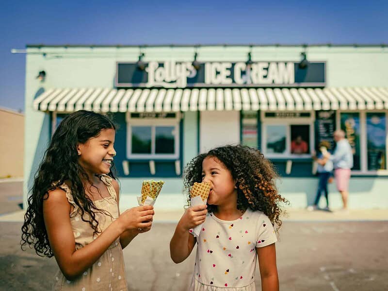 Two young girls stand in front of Lolly's Ice Cream shop, smiling and holding ice cream cones, with people and a menu board in the background.