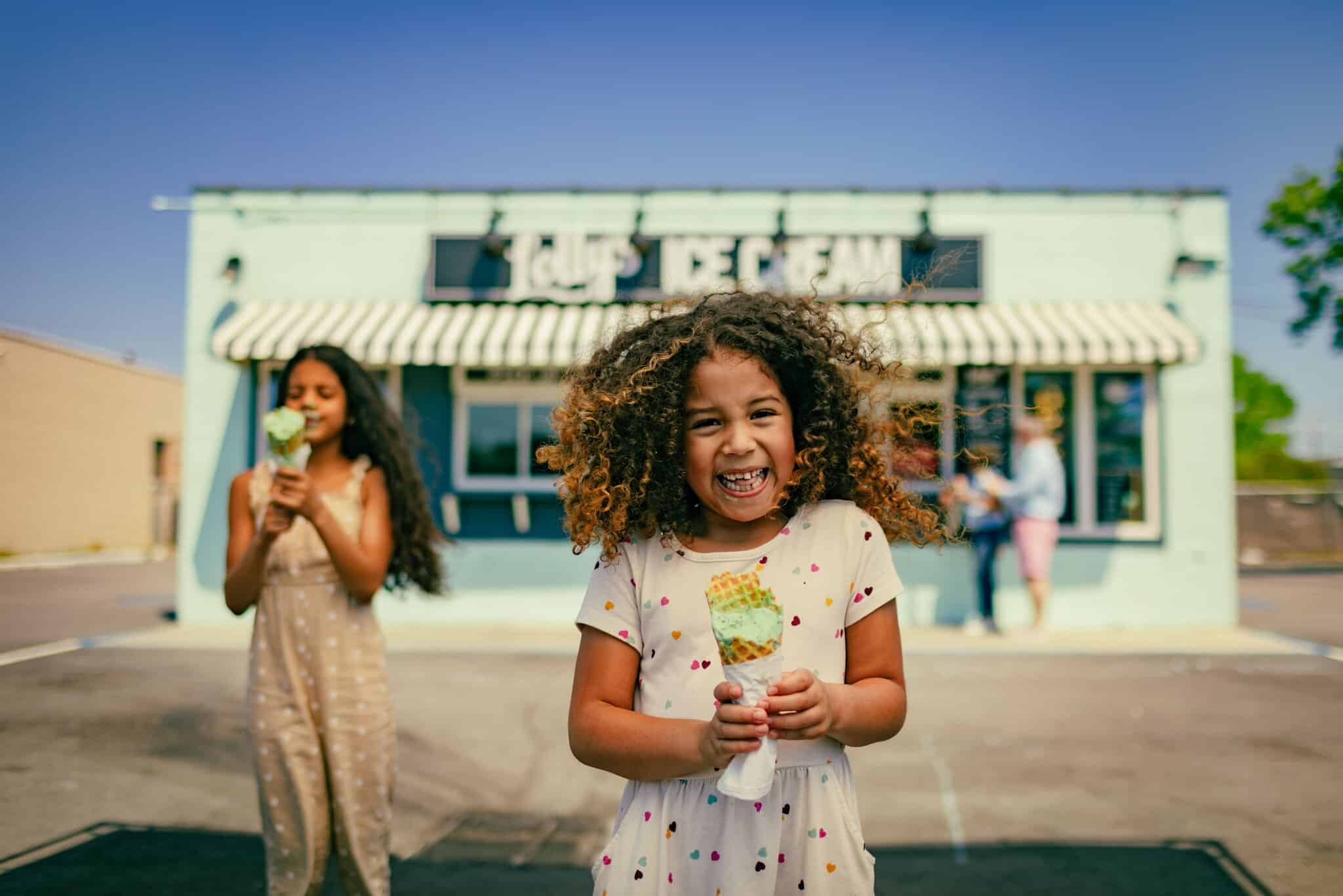 Two children stand outside Lolly's Ice Cream shop on a sunny day, smiling and holding ice cream cones, with the shop and a striped awning in the background.