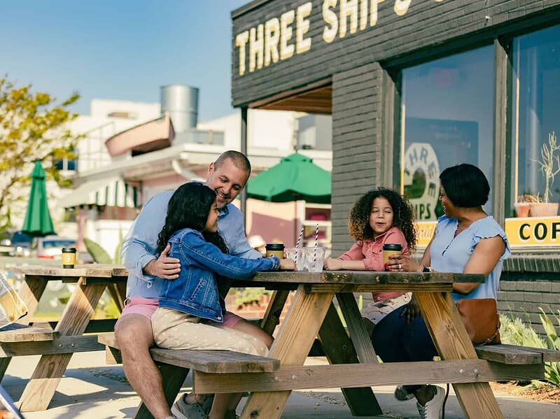 A family of four sits at a wooden picnic table outside of Three Ships Coffee Roasters, drinking beverages and talking on a sunny day.