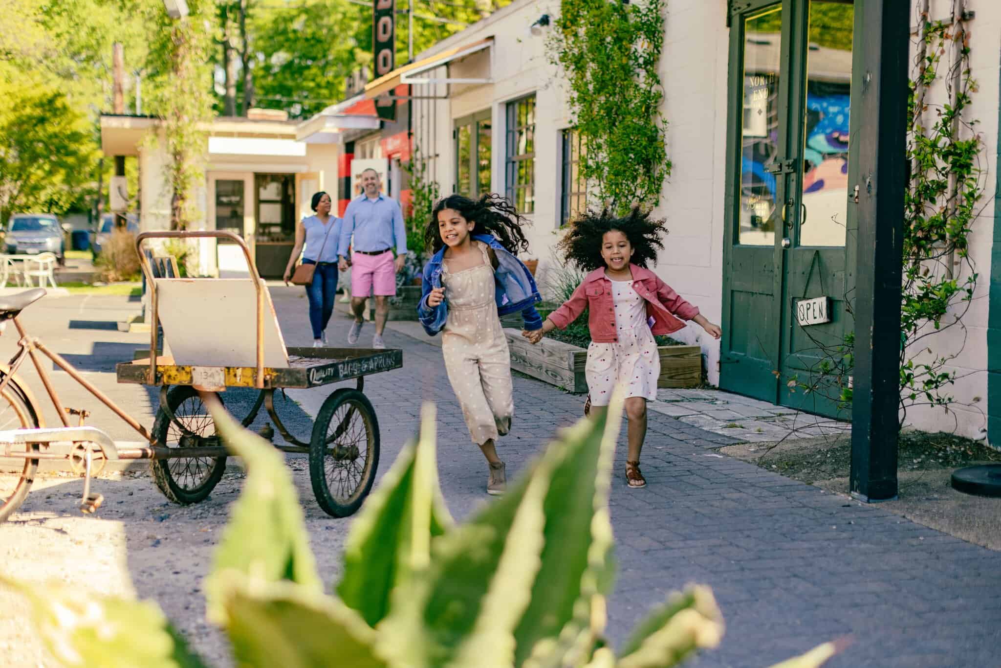 Two girls run and smile on a sidewalk in front of shops while a group of adults walk in the background. A wheelbarrow and plants are also visible.