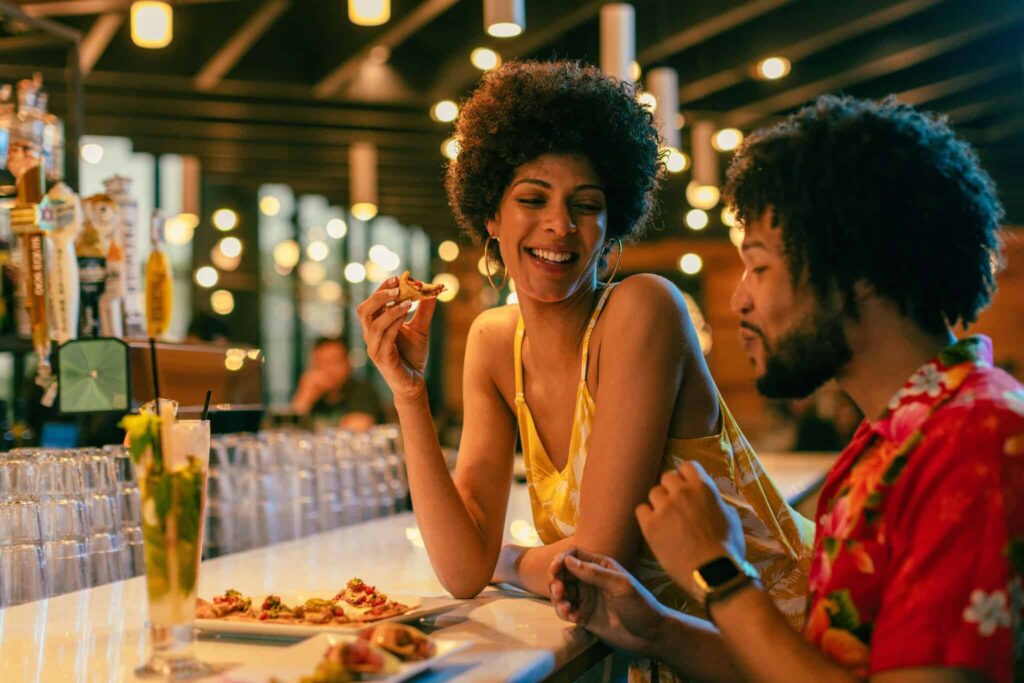 Two people sit at the bar at Cantina Laredo in Virginia Beach enjoying food and drinks, chatting and smiling under warm lights.