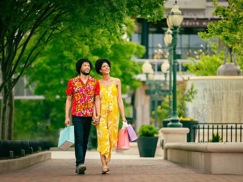 A couple walks outdoors on a brick path at the Town Center, holding shopping bags and hands, surrounded by trees, street lamps, and a large fountain in the background.