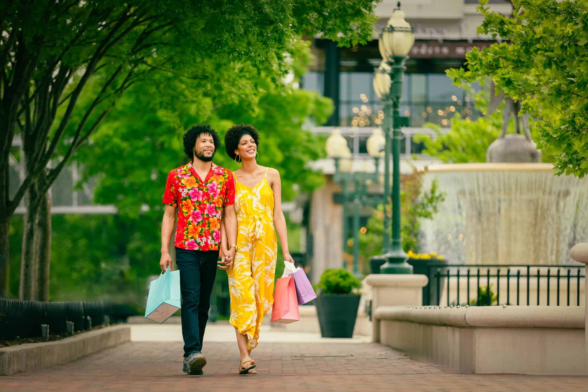 A couple walks outdoors on a brick path at the Town Center, holding shopping bags and hands, surrounded by trees, street lamps, and a large fountain in the background.