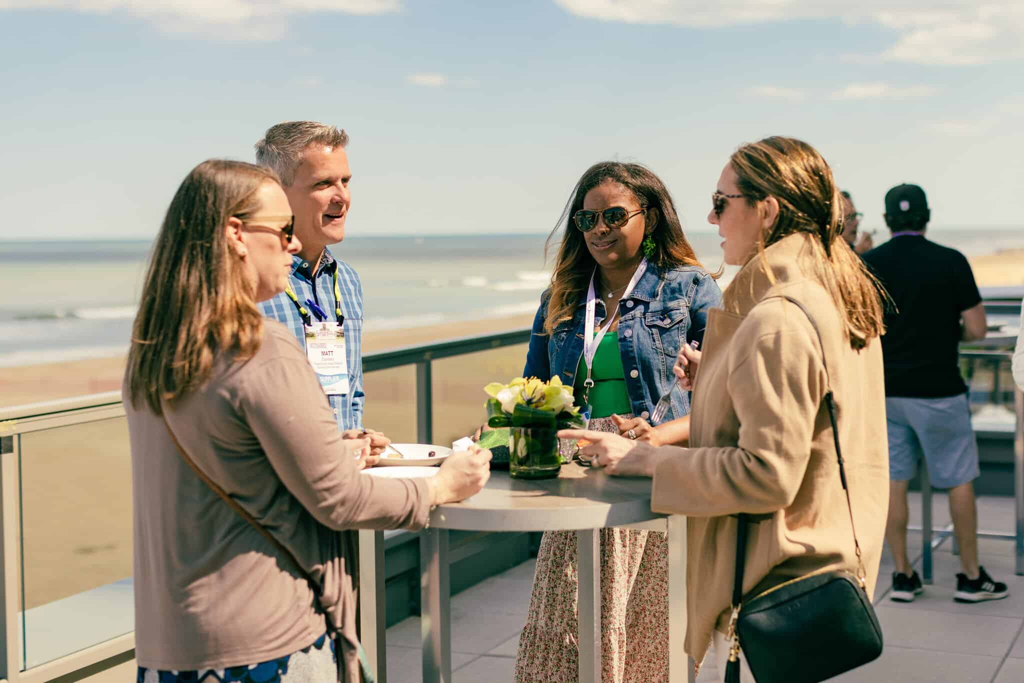 Four people stand around a tall table outdoors near a beach, talking and smiling. Two women wear sunglasses, and a potted plant sits on the table.