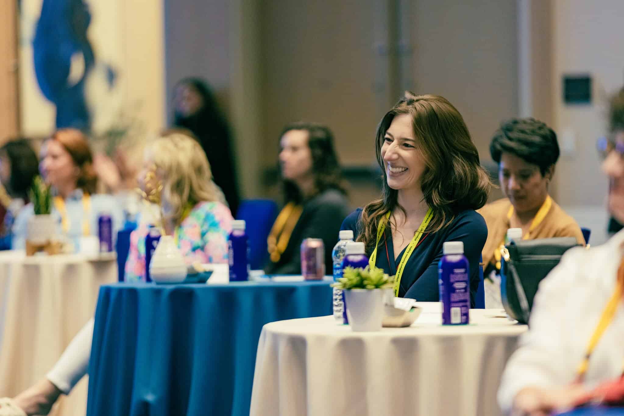 A group of people sit at round tables with water bottles and notepads, attentively listening during an indoor conference or seminar at the Marriott Hotel.