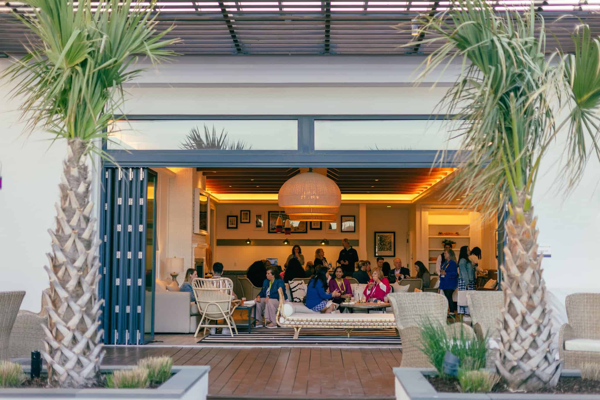 A group of people are gathered inside a modern open-air lounge, viewed from a patio with palm trees and wicker furniture in the foreground.