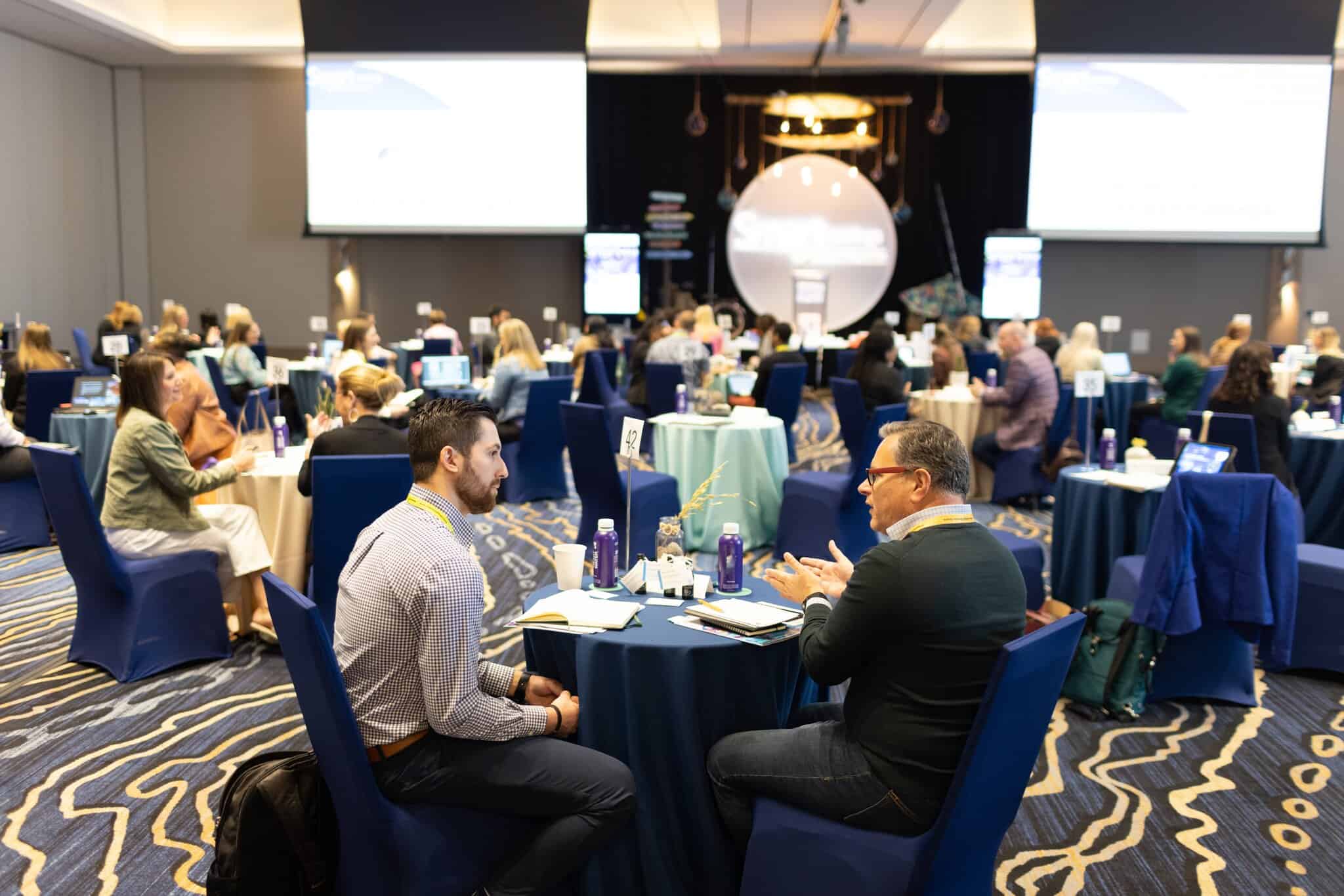 People sit at round tables in a conference room having discussions, with presentation screens and event materials visible in the background.