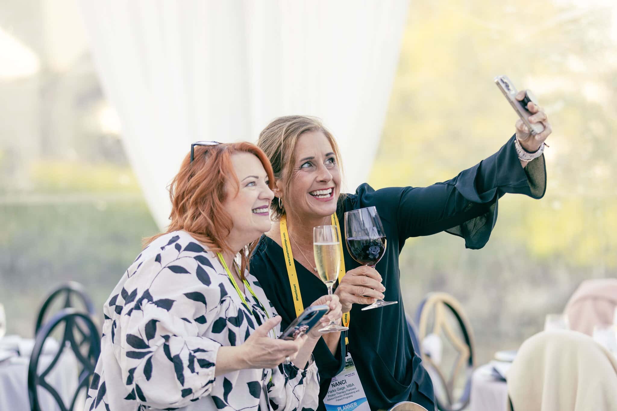 Two women stand together, smiling and holding drinks, as one of them takes a selfie with a smartphone at a formal event.