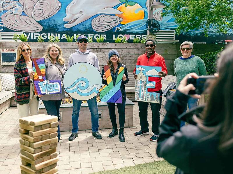 Six people stand outdoors holding colorful letters spelling "VIBE" as another person takes their photo; a mural and trees are in the background.