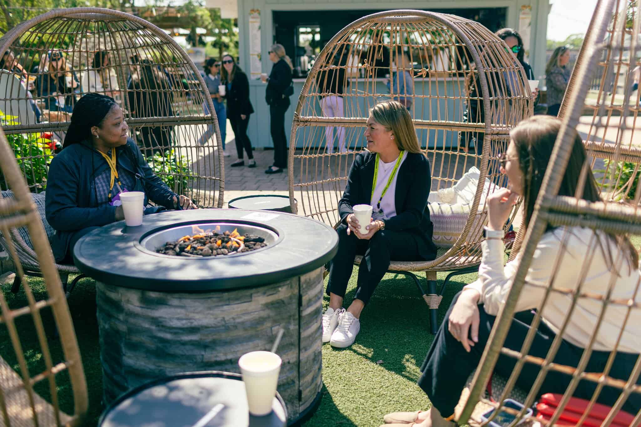 Three women sit around a circular outdoor fire pit, talking and holding cups, with others and greenery visible in the background.