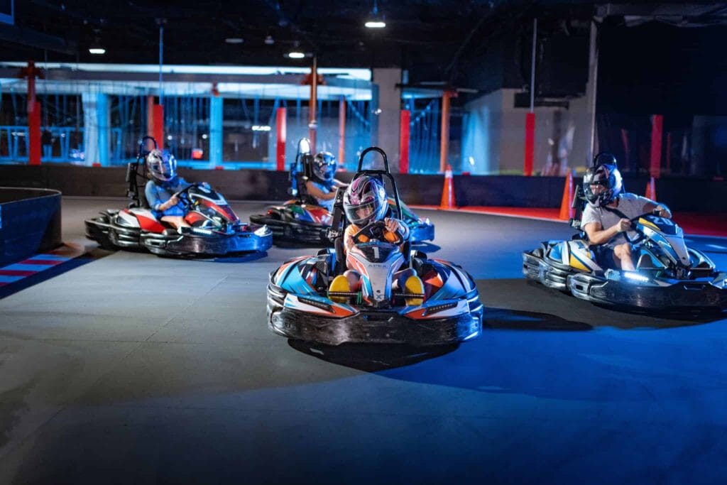 Four people wearing helmets drive go-karts on an indoor track at Apex Entertainment, each in their own vehicle, with barriers and orange cones visible in the background.