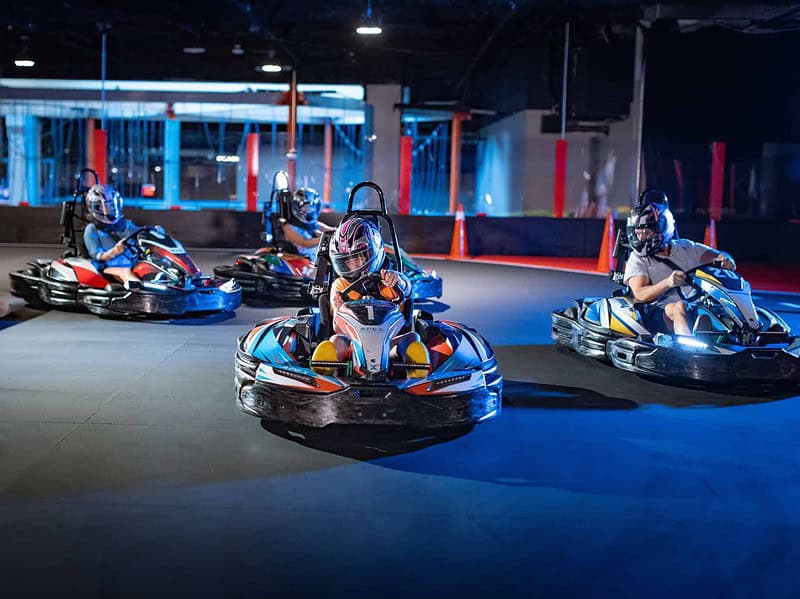 Four people wearing helmets drive go-karts on an indoor track at Apex Entertainment, each in their own vehicle, with barriers and orange cones visible in the background.