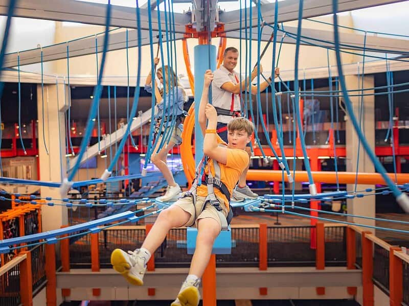 A boy in a harness navigates a rope course while two adults follow behind him. The indoor area features bright orange and blue structures.
