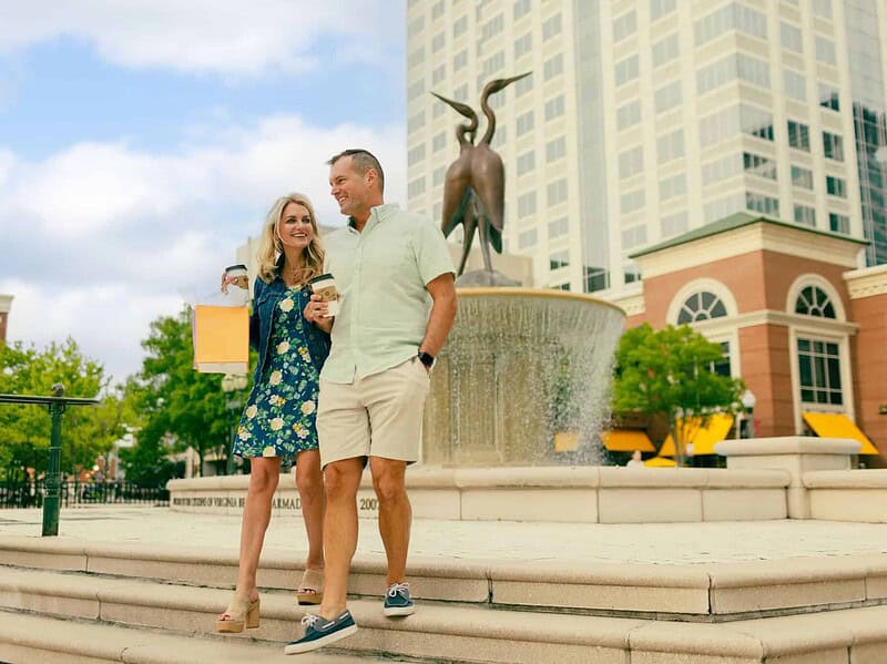 A man and woman walk down steps holding hands and shopping bags near a fountain and tall building in an urban area on a sunny day.