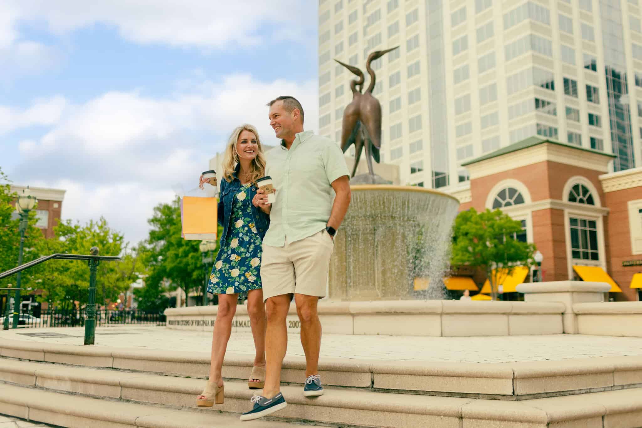 A man and woman walk down steps holding hands and shopping bags near a fountain and tall building in an urban area on a sunny day.