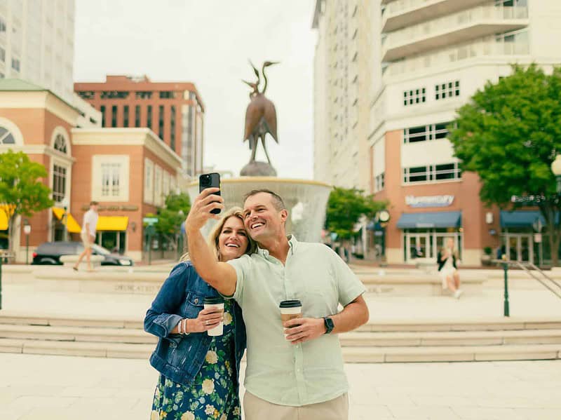 A smiling man and woman take a selfie together while holding coffee cups in front of an outdoor fountain at the Town Center.