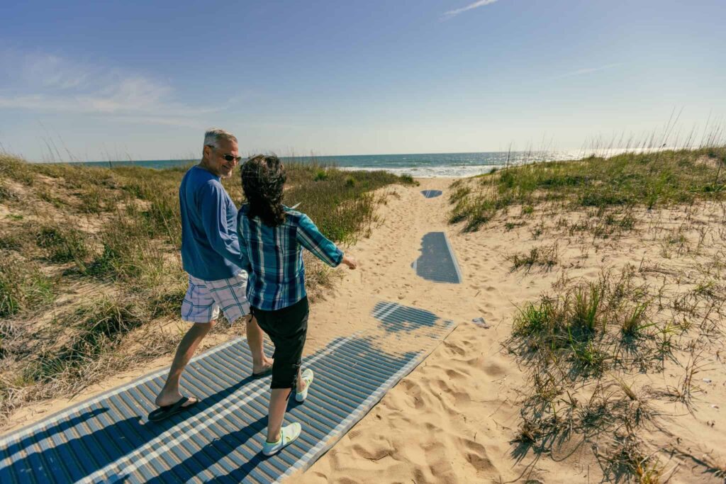 Two people walk together on a mat-covered path through sandy dunes toward the ocean on a sunny day at Back Bay.