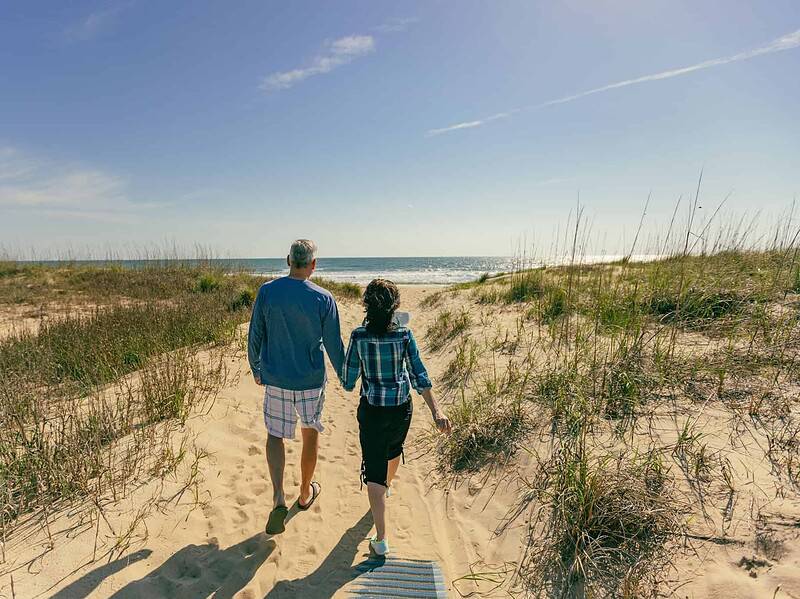 Two people walk hand in hand on a sandy path at Back Bay through dune grass toward the ocean under a clear blue sky.