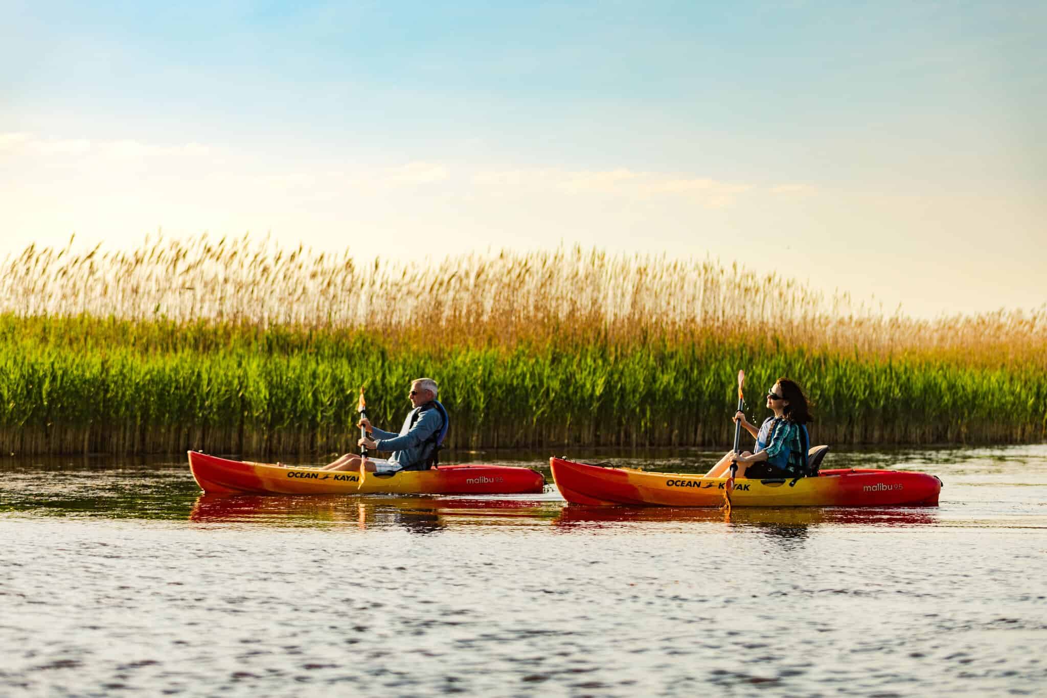 Two people paddle orange kayaks at Little Island Park with tall grass and blue sky in the background.