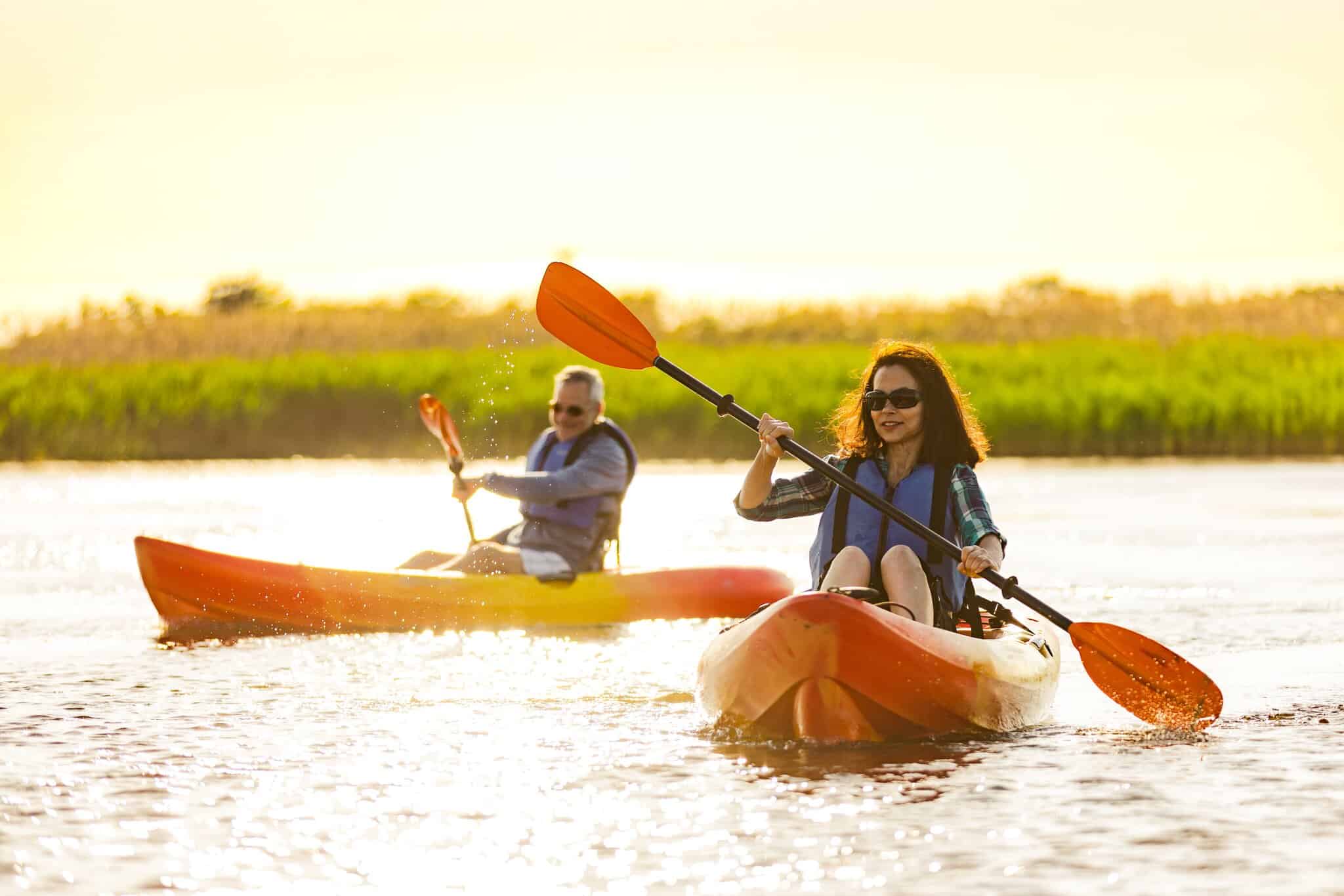 Two people wearing life jackets paddle kayaks at Little Island Park with tall grass visible in the background.