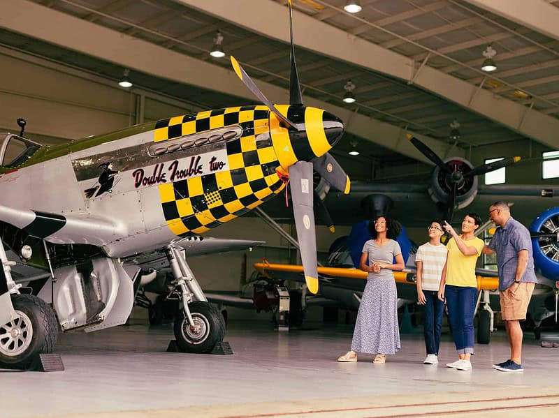 Four people stand near a vintage airplane with a yellow and black checkered nose inside an aircraft hangar, observing and discussing the exhibit.