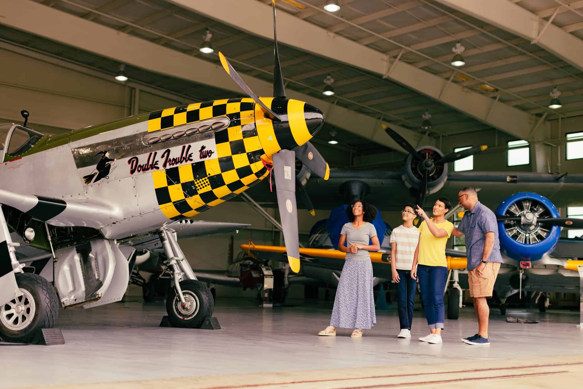 Four people stand near a vintage airplane with a yellow and black checkered nose inside an aircraft hangar, observing and discussing the exhibit.