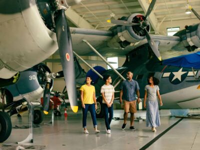 A family of four walk together inside an aircraft hangar at the Military Aviation Museum, with large vintage military planes in the background.